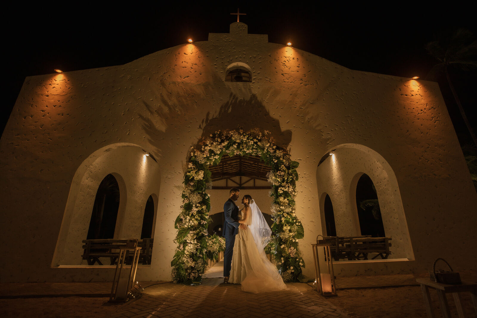 Thalita e Léo Medina sob o arco floral da capela iluminada, à noite.